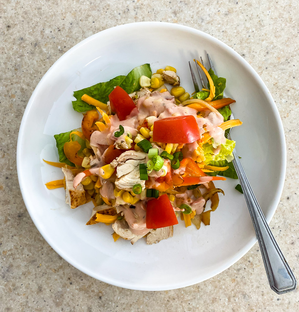 Mix ranch and salsa together to make the dressing, set aside. Rest chicken for 10 minutes then chop. Layer lettuce in a large bowl or serving platter.  Add the peppers and onions (and yummy juices), chicken pieces, corn, cheese, tomatoes and the green onion. Top with some dressing!